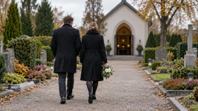 Zwei Trauergäste gehen mit weißen Blumen auf einem Friedhof zur Trauerhalle.